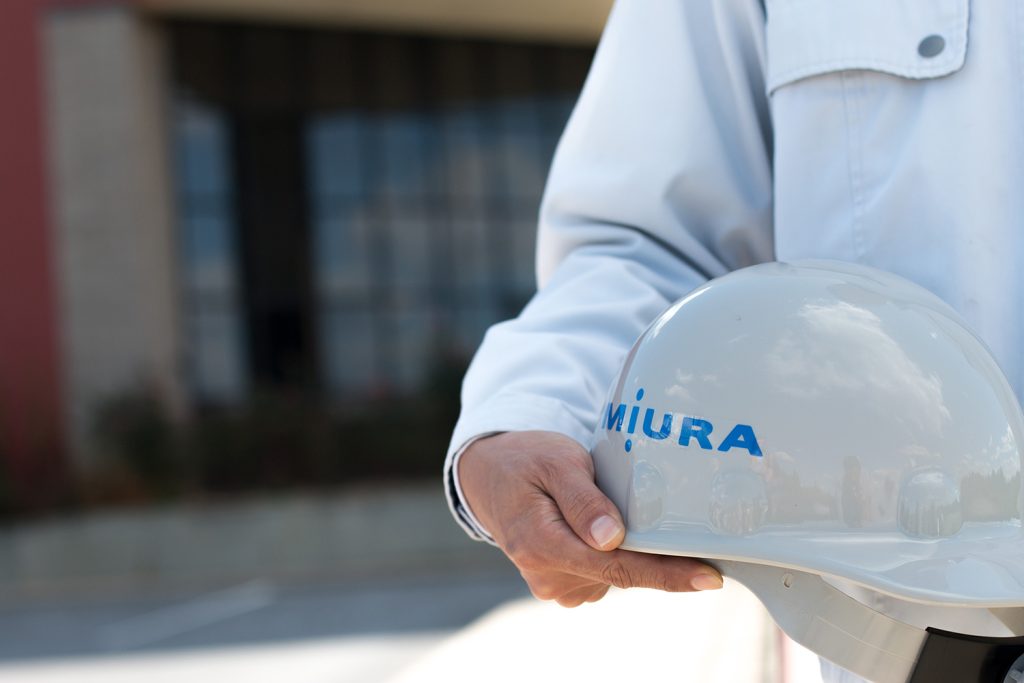 Miura Canada safety image, close up of a man in grey jacket holding a white Miura hard hat.